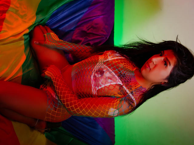 IsabellaStonnee with red fishnet outfit, long black hair, red face paint, sitting on colorful rainbow fabric, looking at camera, red and green lighting, medium-sized tits, cute, sexy.