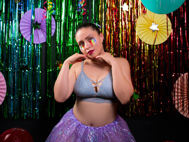 AliceBlackx, con il trucco colorato sul viso, un reggiseno a fascia con lacci blu e una gonna a balze viola scintillante, con le mani vicino al mento, guarda la fotocamera. Ha un seno grande e un sedere prosperoso..