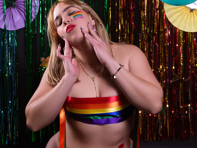 ZaharaBennet with rainbow makeup, hands on face, wearing a rainbow-striped bandeau covering her tits, colorful ribbons in background, red lipstick, gold necklace, and purple nails.