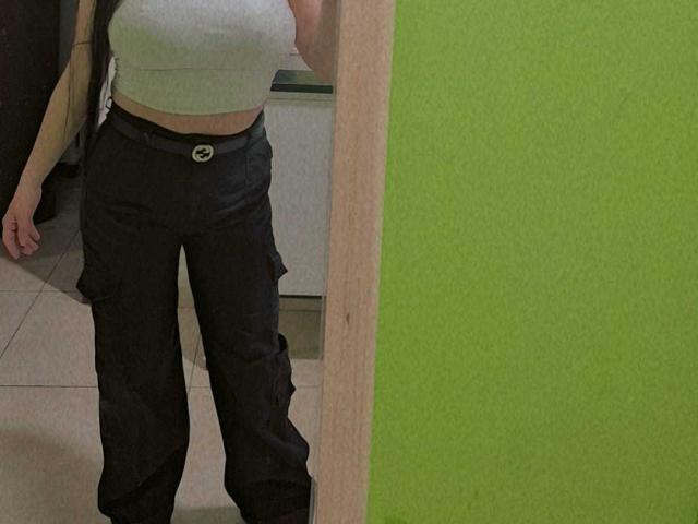 HoneyHotX mit einem schlanken, geschwungenen Körper, trägt ein eng anliegendes, graues Crop-Top, schwarze Hosen und einen sichtbaren Gürtel und steht in einem grün gestrichenen Raum und zeigt ihr kleines, festes Hinterteil..