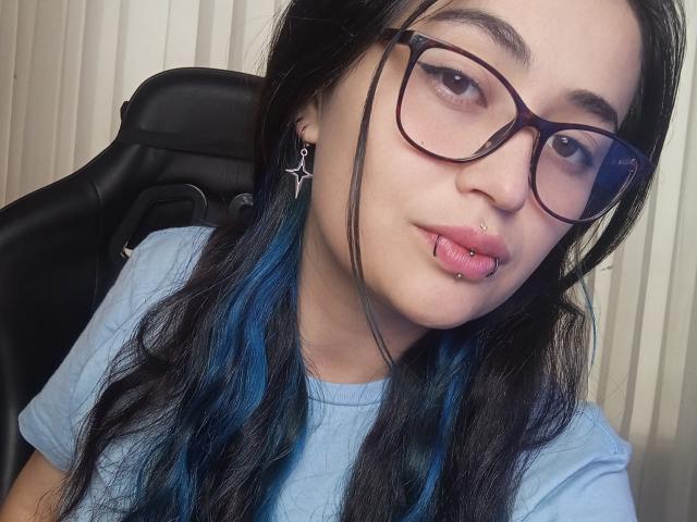 EvaMiller69 met blauw geverfd haar, een bril, een neusring en een lip piercing, draagt een lichtblauwe blouse en zit in een zwarte stoel, kijkend naar de camera..