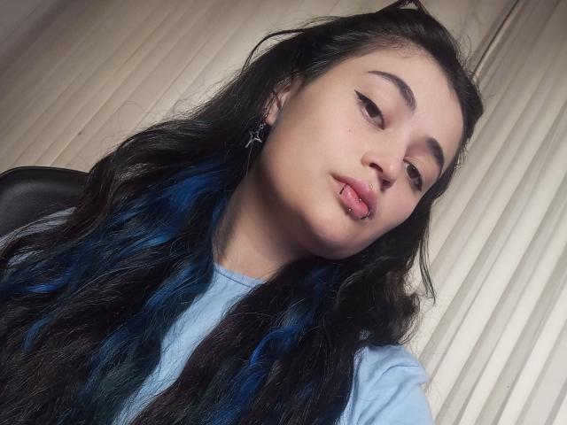 EvaMiller69, con cabello largo negro y azul, piel clara, cejas pobladas y la lengua afuera, vestida con una camisa azul claro, en un selfie. Sus senos no son visibles..