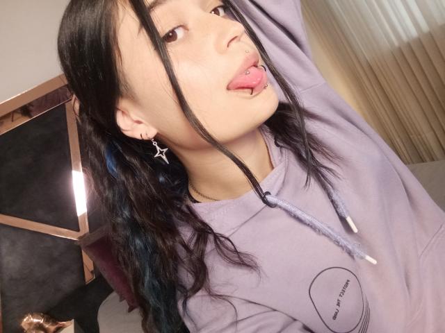 EvaMiller69, avec des cheveux noirs, la langue tirée, un sweat à capuche violet, un anneau au nez et des boucles d'oreilles en forme d'étoile, la langue tirée avec un piercing en forme de bille rose, regardant la caméra..