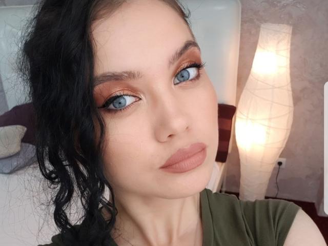 KimHonney with blue eyes, heavy eyeliner, and wavy black hair, wearing green, has full pink lips and a serious expression. Her tits are not visible.