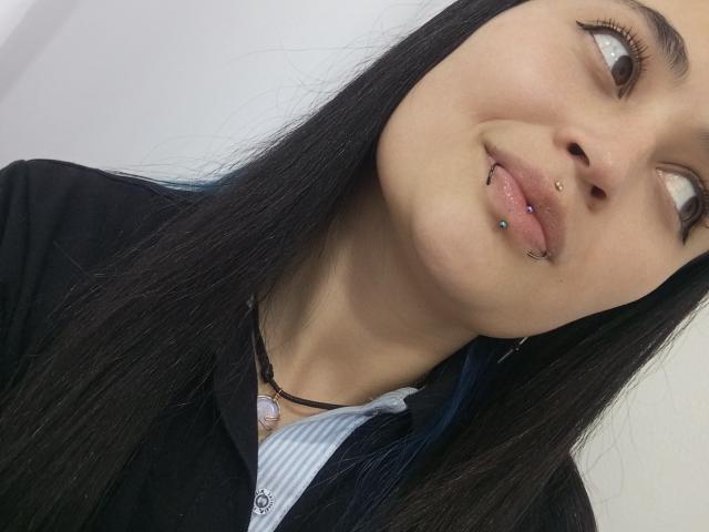 EvaMiller69 with long black hair, upturned nose, and multiple piercings (lip and nose), wearing a black shirt over a white striped top, looking to the side.