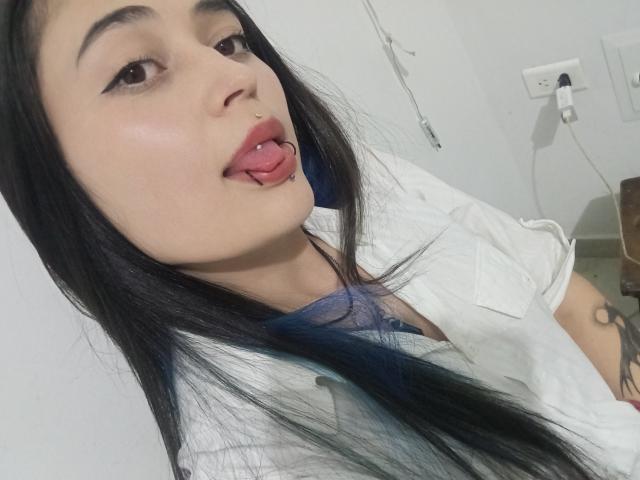 EvaMiller69 mit langem, schwarzem Haar, blauen Spitzen, einem weißen Hemd und einem Nasenring, die ihre Zunge herausstreckt (mit einem Zungentattoo) und ihre kleinen Brüste zeigt..