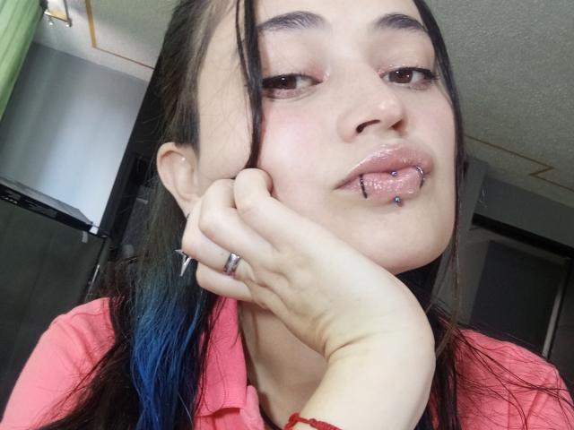 EvaMiller69 tiene el cabello teñido de azul, ojos marrones y un piercing en el labio plateado con una bolita azul. Lleva una camisa rosa, se sostiene la barbilla y está mirando a la cámara..