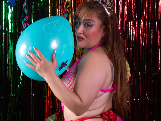 MeryLand, con un bikini a rete rosa, tiene in mano un palloncino blu, ha il rossetto rosso, l'ombretto blu e i capelli castani lunghi, indossa una tiara d'argento, mostrando i suoi fianchi prosperosi e il seno grande..