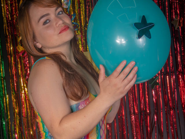 MoonRay69 met lichtbruin haar, die een blauwe ballon vasthoudt met een sterstickertje, draagt een kleurrijke, gestreepte jurk, kijkt omhoog met een lichte glimlach en laat haar borsten zien..