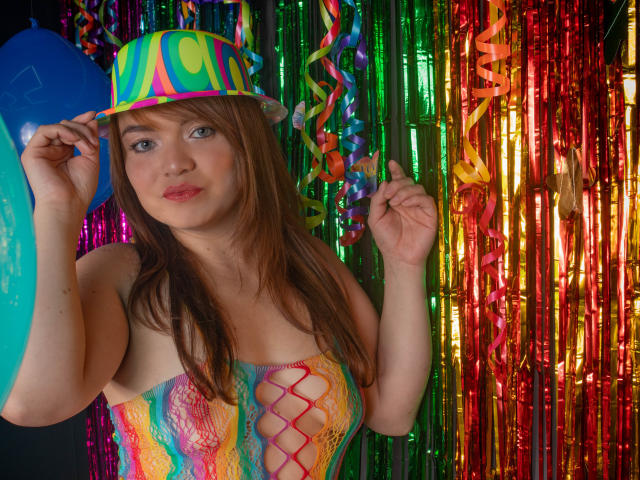 MoonRay69 mit braunen Haaren, die eine Neonmütze trägt, ein buntes Schnürkleid anhat und einen blauen Luftballon hält, hat Brüste, die durch das Kleid sichtbar sind, und steht vor einem glänzenden, bunten Vorhang..