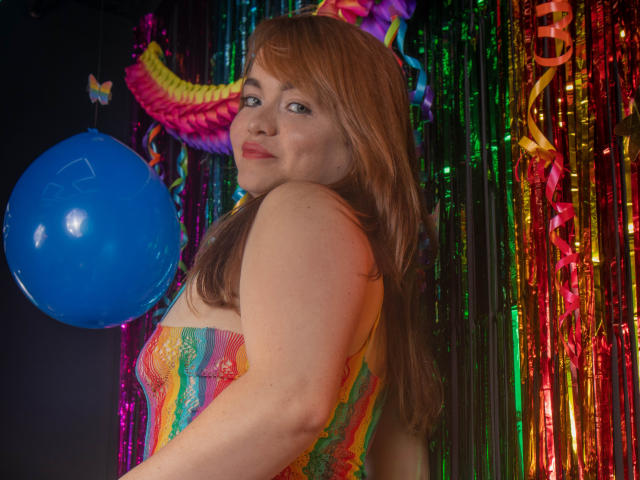 MoonRay69, avec de longs cheveux roux, portant un corset en dentelle coloré, regardant par-dessus son épaule, souriant à la caméra, un ballon bleu à gauche, des guirlandes colorées en arrière-plan..