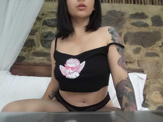JeneKurz com blusa preta que deixa os ombros à mostra, exibindo uma tatuagem de anjo rosa, calcinha de renda preta, tatuagens nos braços, sentada na cama, seios médios, cabelo preto, fundo com parede de pedra..