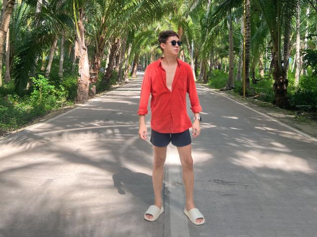 Johnerick with short brown hair, wearing a red button-up shirt, dark shorts, white sandals, and sunglasses, standing on a paved path with tall green palm trees.