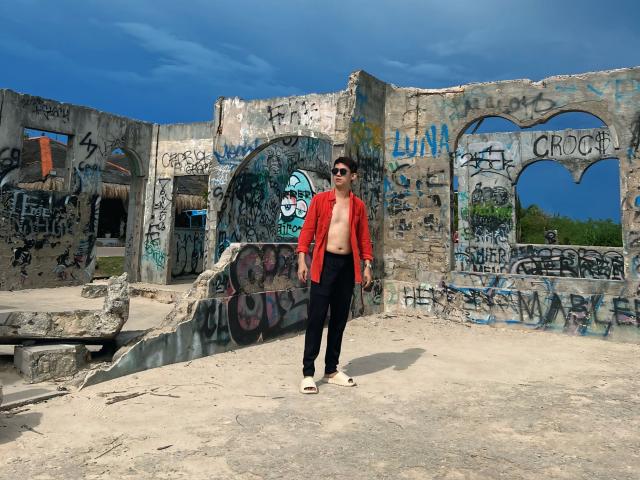 Johnerick stands in front of a graffiti-covered, ruined concrete wall, wearing a red open shirt, black pants, and sandals, with sunglasses on, and a shirtless upper body.