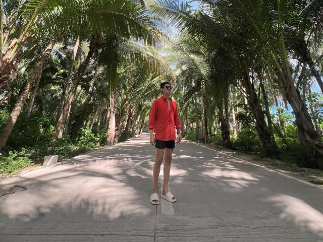Johnerick stands on a sunny path, wearing a red shirt, black shorts, white slip-on shoes, and sunglasses, with tall palm trees in the background.
