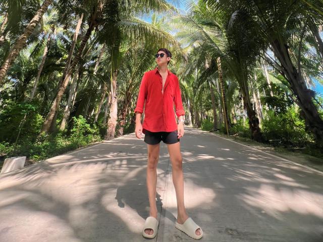Johnerick stands on a sunlit path, wearing a red shirt, black shorts, white sandals, and sunglasses. Tall palm trees surround her. She's slim with long legs.