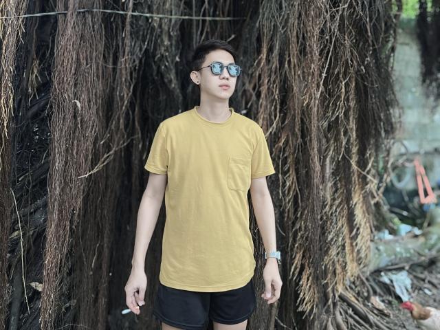 Johnerick with short brown hair, wearing a yellow t-shirt, black shorts, and reflective sunglasses, standing in front of a tree with long, brown, hanging vines. Wearing a silver watch.