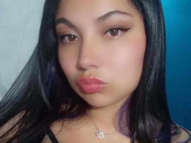 AbbieJones mit sinnlich geschwungenen, rosafarbenen Lippen, braunen Augen und langen, schwarzen Haaren, trägt ein schwarzes Oberteil und eine kleine Halskette mit einem herzförmigen Anhänger..