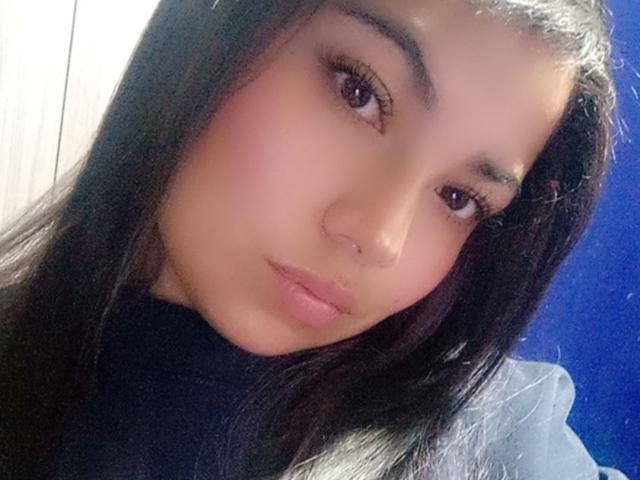 AbbieJoness hat eine mittelbraune Haut, langes schwarzes Haar und volle, rosafarbene Lippen. Sie schaut in die Kamera, wobei ihre rosafarbenen Lippen leicht geöffnet sind und sie ein dezentes Make-up trägt. Ihre Brüste sind nicht sichtbar..