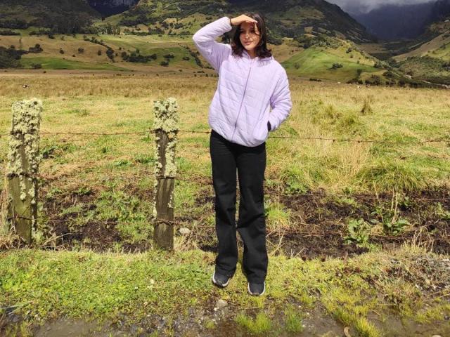 LanaKlaire with medium tits, wearing a purple jacket and black pants, standing in a grassy field, hand in hair, smiling, with a wooden fence and mossy posts.