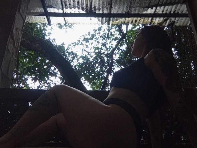 JeneKurz, com tatuagens escuras, um top esportivo preto e calcinha preta, sentada com as pernas dobradas, de costas para a câmera, sob um telhado com folhas de árvores visíveis através dele..