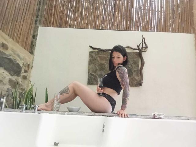 JeneKurz with tattoos, black lingerie, sitting on a white bathtub, legs crossed, showing her ass, looking at camera, bamboo wall in background.
