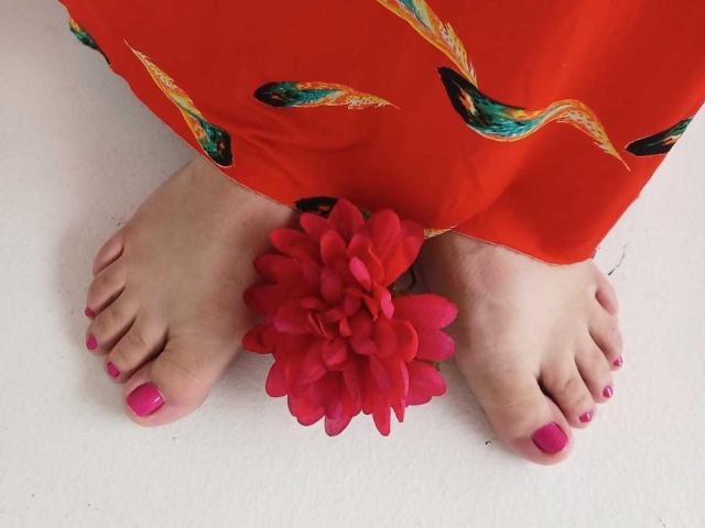 RebeccaSinss's feet, pink-painted toenails, red flower between toes, orange dress with colorful bird pattern, white floor, soft light, slight shadow.