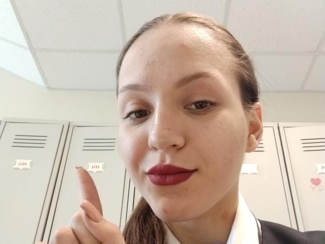 CelieAnnette, with a slight smile and pink lipstick, points a finger up, showing a confident gesture, in a locker room with gray metal lockers in the background.