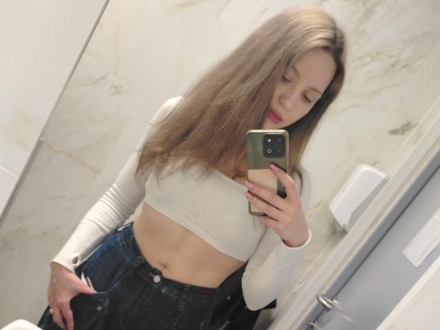 CelieAnnette with long brown hair, white crop top, and blue jeans, showing her flat stomach and hand in her jeans, taking a mirror selfie with a green phone.