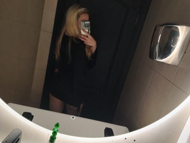 CelieAnnette with long blonde hair, black top, and no pants, taking a mirror selfie in a bathroom. Her phone covers her face. Green bottle on the sink.