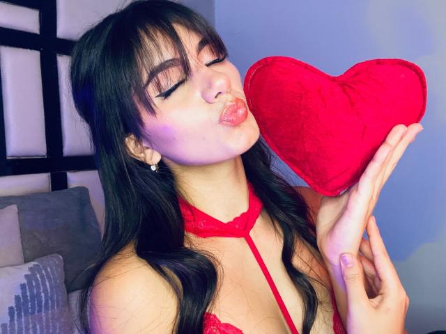 AndreaKerry with black hair, closed eyes, and puffy lips, kissing a red heart-shaped pillow, wearing red lace lingerie, holding it to her face, in a bedroom.