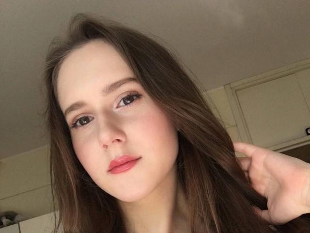 LucyRosen has light skin, brown eyes, and brown hair, looking at the camera with pink lips, holding her hair with one hand, showing a slight smile.