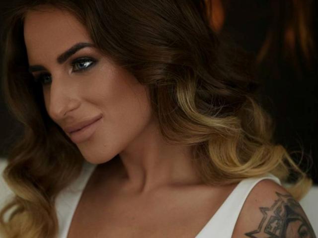 LaymaBlast tiene el cabello ondulado, de color marrón a rubio, maquillaje de ojos ahumado y una pequeña sonrisa. Lleva una blusa blanca que deja ver un tatuaje en su hombro derecho..