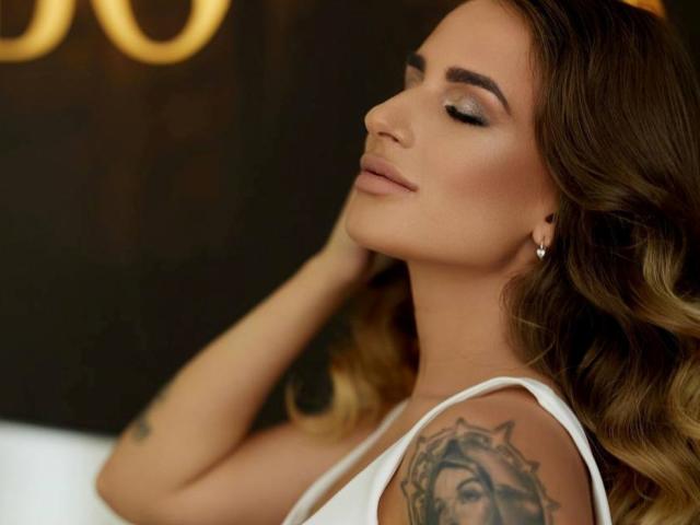 LaymaBlast, com os olhos fechados, cabelo castanho ondulado, blusa branca, tatuagem no ombro direito e brincos de diamante, tocando o pescoço. O tamanho dos seios é médio, a pele é bronzeada..