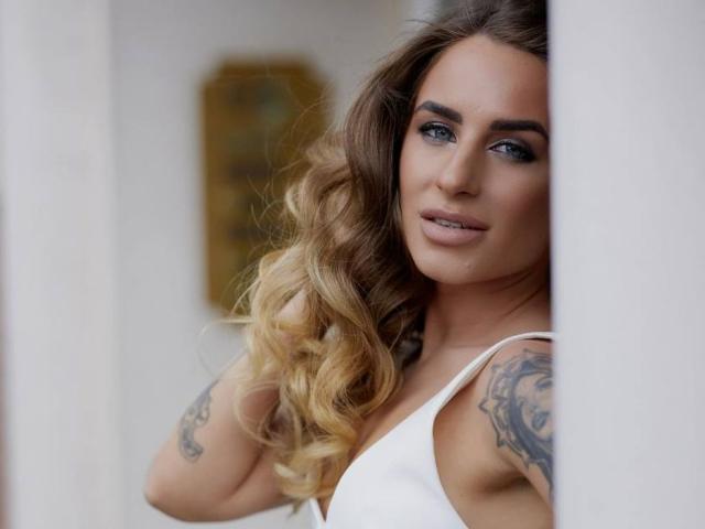 LaymaBlast with wavy blonde hair, tattoos on both arms, and blue eyes, wearing a white dress, standing partially behind a white column, looking seductively at the camera.