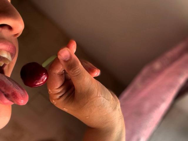La lingua umida di Madrinia sporge, leccando una ciliegia rossa e lucida tenuta nella sua mano sudata. La sua bocca è leggermente aperta, mostrando i suoi denti. La sua pelle è scura..