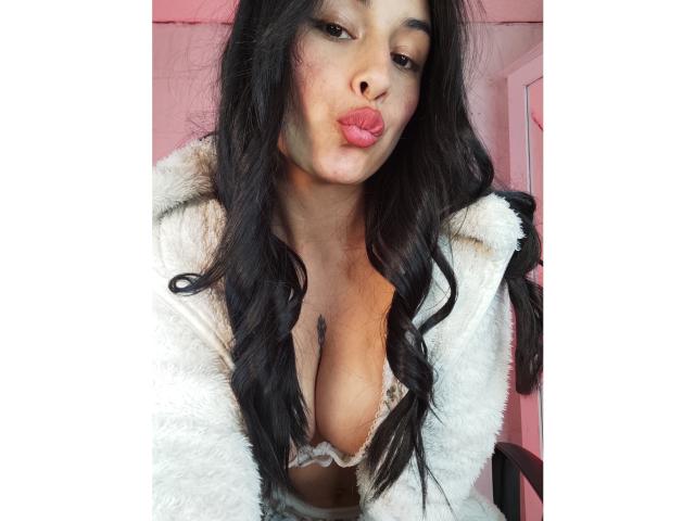 EmilyVv with long black hair, pouty pink lips, wearing a fluffy white jacket, and a lace bra that reveals her big, round, shiny tits.