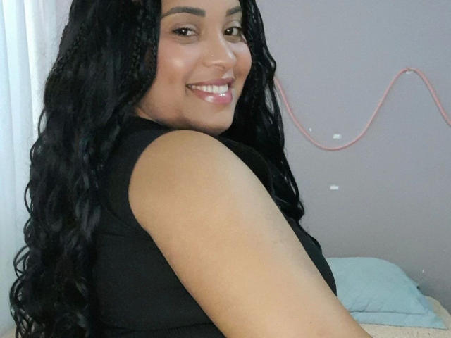 SuelenBarbar4-hot has long black curly hair, a cute nose piercing, and a smile showing white teeth. She's wearing a black sleeveless top, looking back over her shoulder.