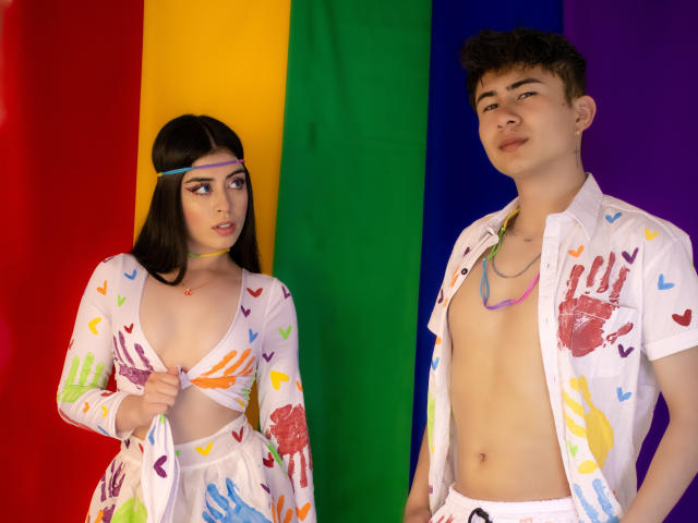 NoahAndRose with dark hair, wearing a colorful handprint dress, showing tits, and a headband, stands next to NoahAndRose with brown hair, shirt open, exposing a cock, wearing white pants.