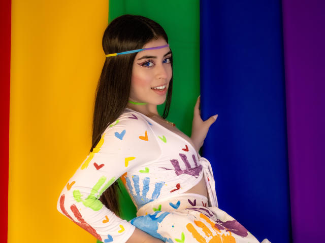 Noah e Rose, com longos cabelos castanhos, vestindo um vestido branco com estampas coloridas de mãos, em pé contra um fundo azul, verde, amarelo e roxo, sorrindo, com delineador azul, usando uma tiara roxa..