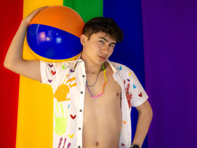 NoahAndRose, shirtless, wearing a white button-up with colorful heart and handprint patterns, holding a rainbow ball, and sporting a multicolored necklace, has short brown hair and a slight smile.