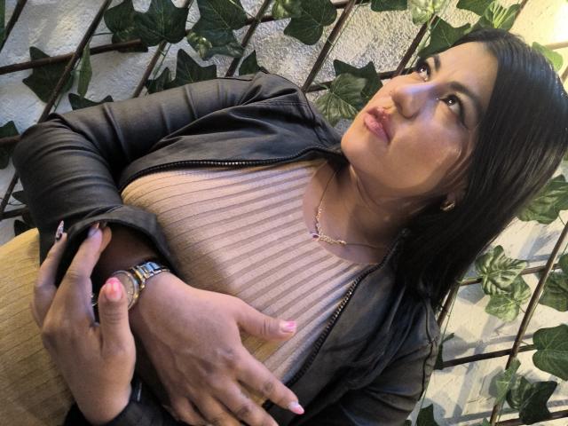 AmaiaCruz with medium tits, wearing a black leather jacket, pink ribbed top, and gold necklace, looking up with pouty lips, hands on chest, green leaf vines in background.