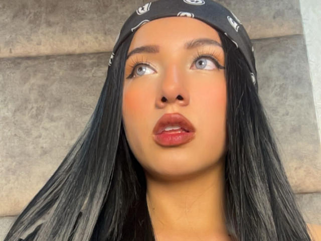 LillyTaylor has dark black hair, blue eyes, and red lipstick. She's wearing a black bandana with white patterns and looking up with a surprised expression.
