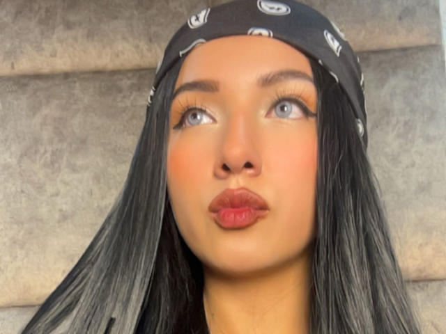 LillyTaylor has puffy ass-colored lips, blue eyes looking up, black headband with white patterns, and long straight black hair, against a grey stone background.