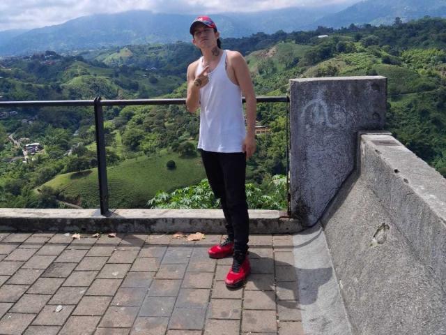 RossAndJack, portant un débardeur blanc, un pantalon noir, des chaussures rouges et une casquette rouge, se tient sur une corniche en briques, tenant un téléphone, avec un mur de pierre en arrière-plan..