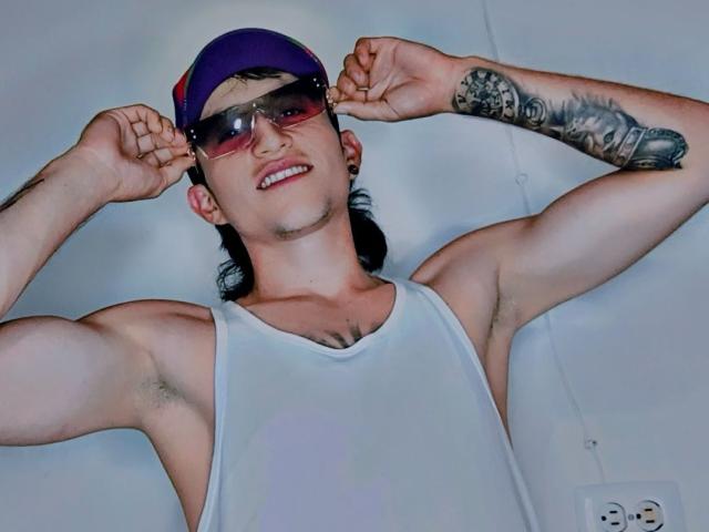 RossAndJack, un uomo muscoloso con una canotta bianca, un berretto viola e occhiali da sole, che mostra i bicipiti, con un braccio destro tatuato, sorridente, con peli visibili sotto le ascelle e pochi peli sul petto..