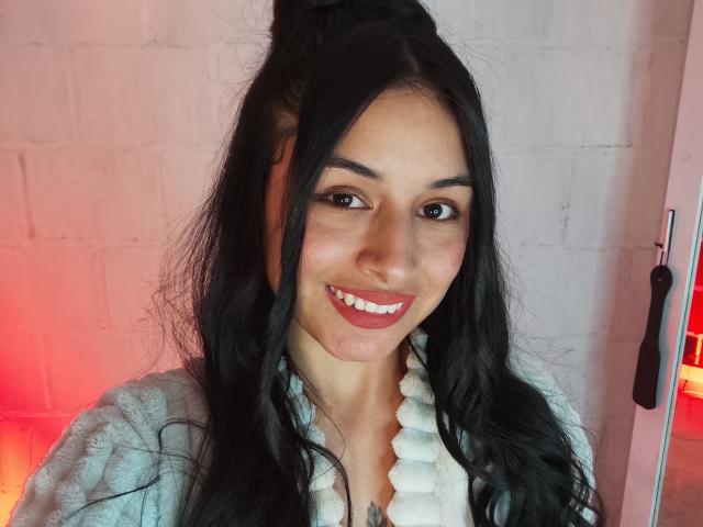 EmilyVv has long black hair, brown eyes, and a cute smile with red lipstick. She's wearing a white, fluffy collar shirt and has a small tattoo on her chest.