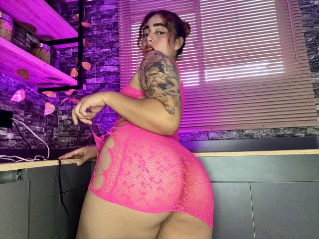 MerydaBraveI with big ass, wearing pink lace lingerie, exposing her butt, tattooed arm, and red lipstick, holding lingerie strings, standing in a room with purple lights.