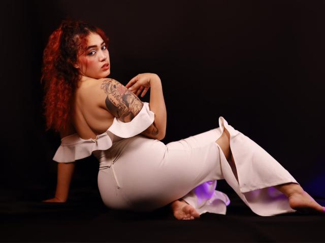 MerydaBraveI has curly red hair, a tattooed arm, and is wearing a white, off-shoulder dress, sitting with her back to the camera, showing her ass.