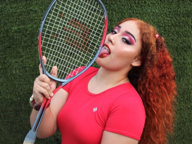 MerydaBraveI with curly red hair, pink eyeshadow, tongue out, holding a tennis racket, wearing a tight red shirt, showing her big tits, against a green grassy background.